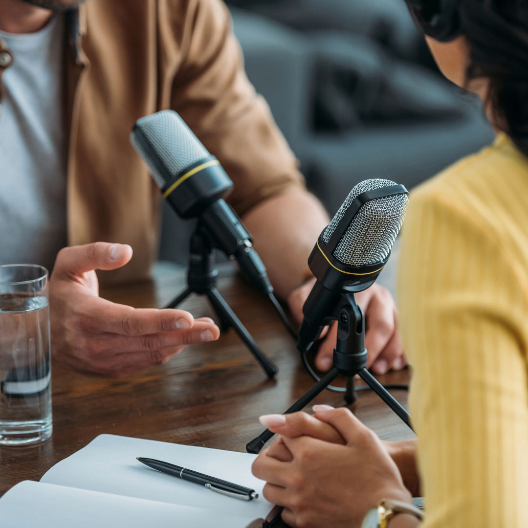 a male and female sitting behind microphones for a podcast